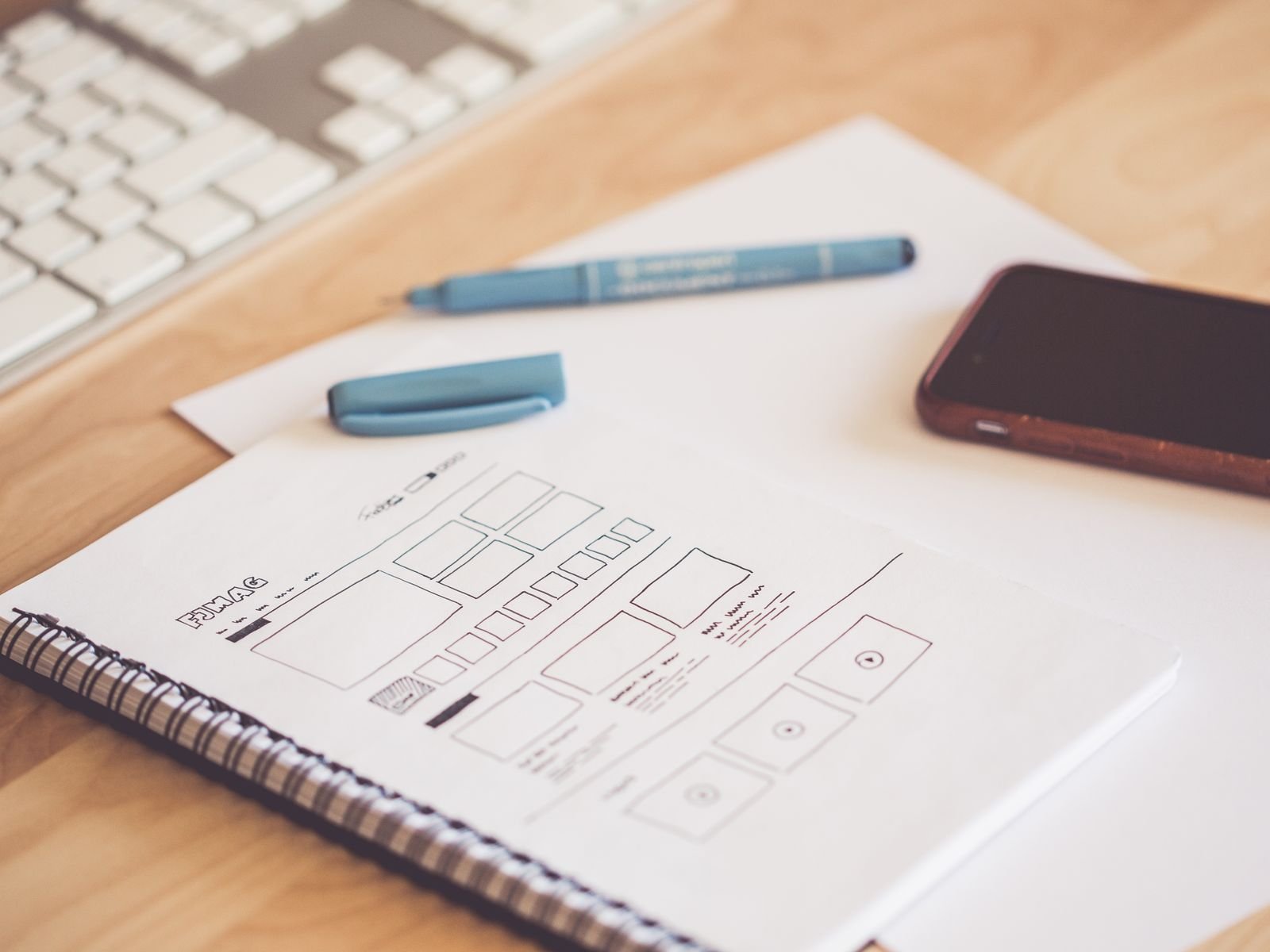 A neat workspace featuring a sketchpad with wireframes, a smartphone, and a keyboard on a wooden desk.